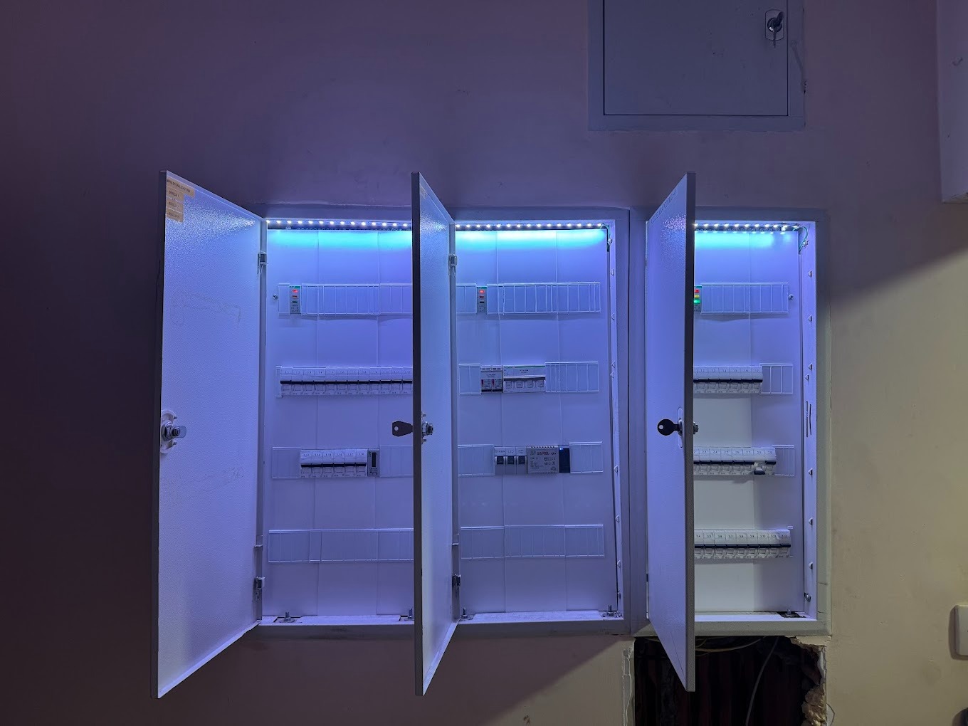 Otwarta, podświetlona listwą LED, trójdzielna skrzynka elektryczna z bezpiecznikami, zamontowana na ścianie z widocznym otworem instalacyjnym w dolnej części.