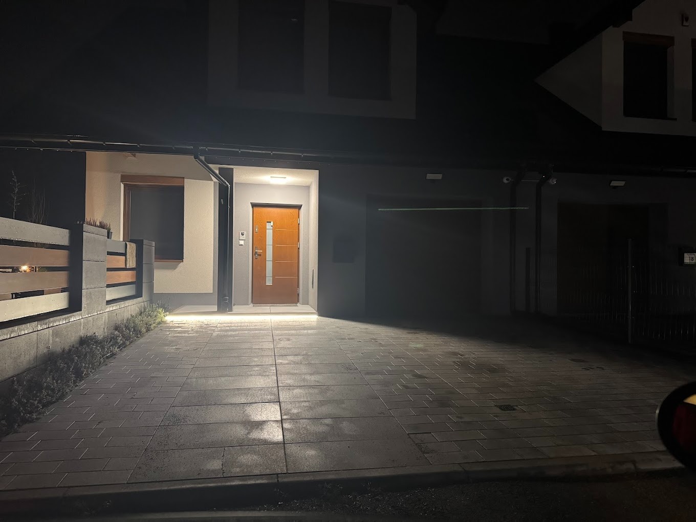 Nocny widok na dom z zainstalowanym oświetleniem zewnętrznym podkreślającym wejście i podjazd wyłożony kostką brukową. Widoczne kamery monitoringu na elewacji.