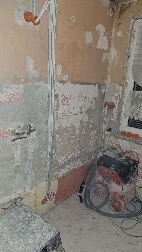 Wnętrze w trakcie remontu z widocznymi przewodami elektrycznymi wyprowadzonymi ze ściany, puszkami instalacyjnymi i odkurzaczem przemysłowym w rogu pomieszczenia.