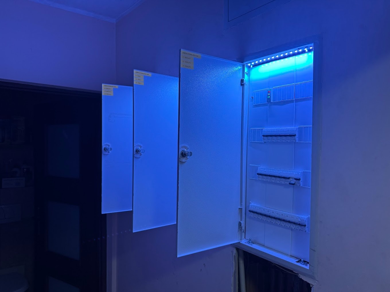 Otwarta skrzynka elektryczna z oświetleniem LED wewnątrz, obok zamknięte skrzynki, oświetlone niebieskim światłem