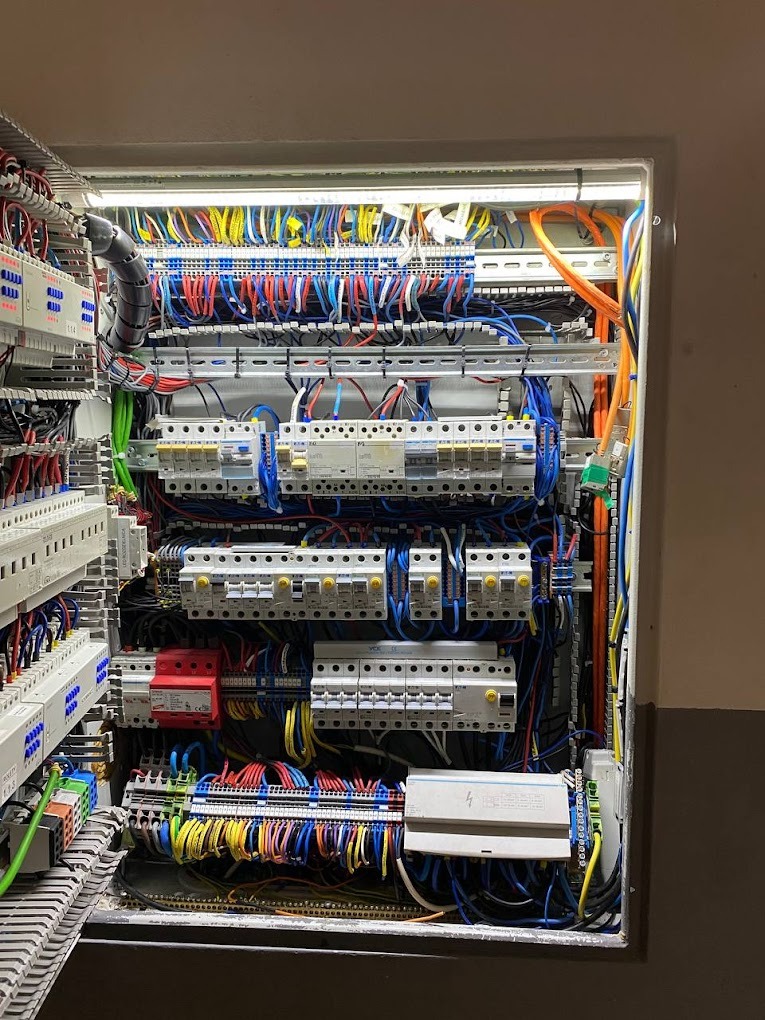 Otwarta skrzynka rozdzielcza z licznymi bezpiecznikami, przewodami w różnych kolorach i elementami mocującymi, oświetlona od góry światłem LED.