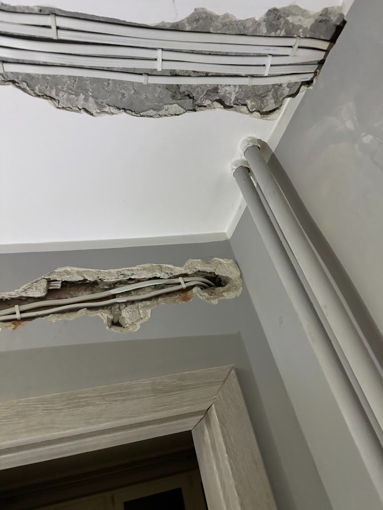 Niedokończona instalacja elektryczna w suficie i ścianie, widoczne przewody zabezpieczone opaskami, fragment rury i ościeżnicy drzwi w szarym pomieszczeniu.