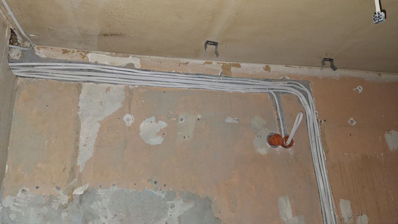 Ściana w trakcie remontu z poprowadzonymi przewodami elektrycznymi w listwach, widoczne puszki instalacyjne w kolorze pomarańczowym, surowy tynk i fragmenty starej farby.