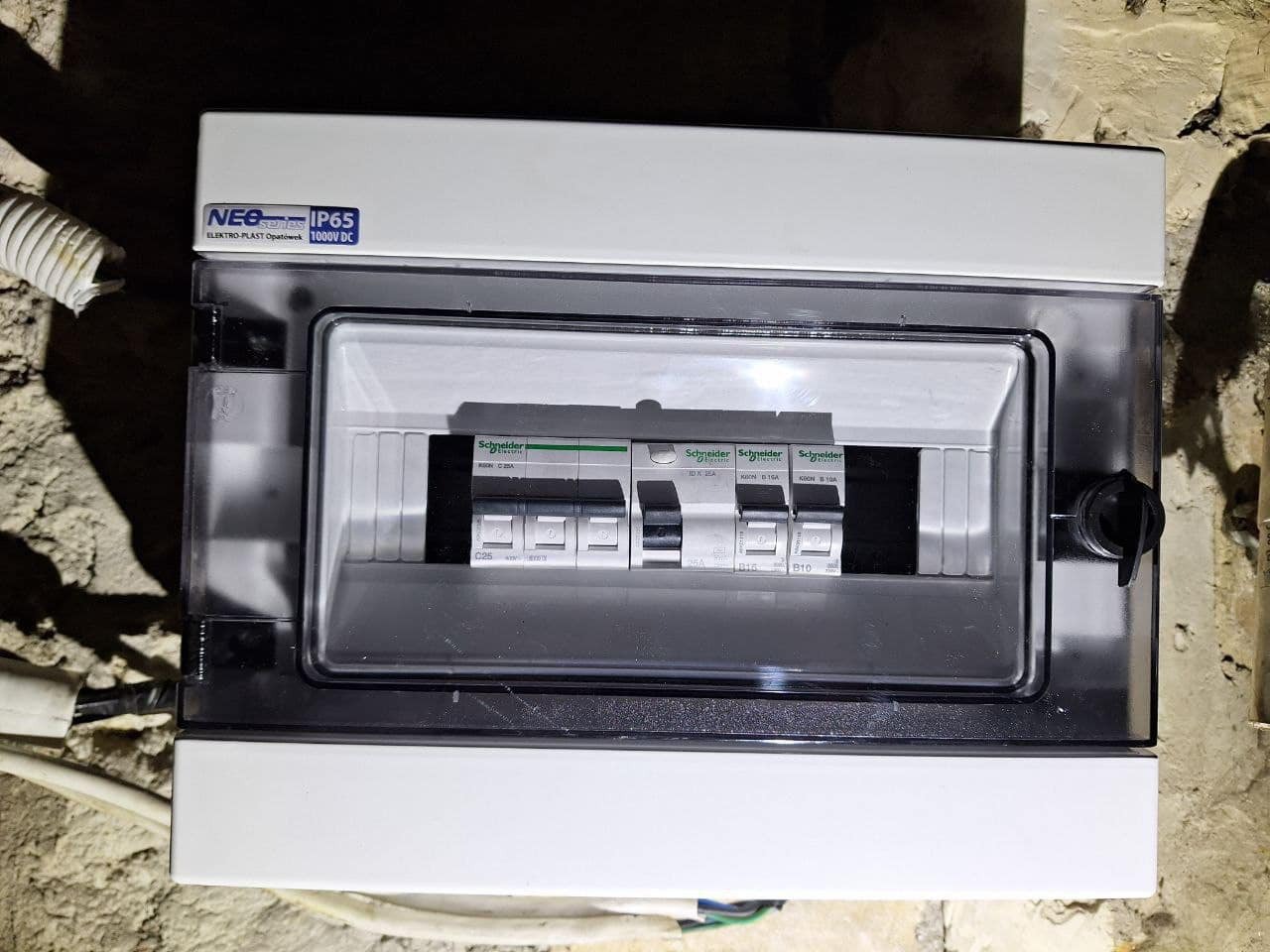 Zamknięta skrzynka elektryczna z widocznymi bezpiecznikami marki Schneider, zamontowana na szorstkiej ścianie. Widoczny przewód ochronny.