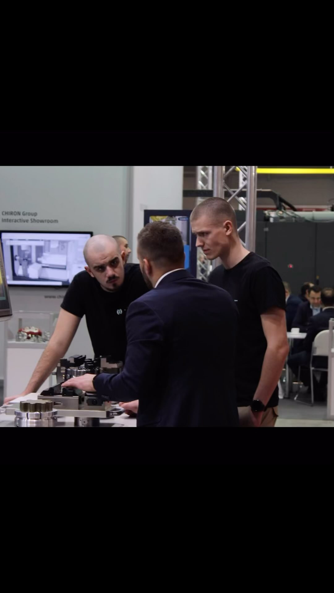 Trzech mężczyzn w pomieszczeniu, jeden w garniturze, dwaj w czarnych koszulkach, pochyleni nad metalowym urządzeniem na stole, w tle ekran z grafiką i logo Chiron Group Interactive Showroom.