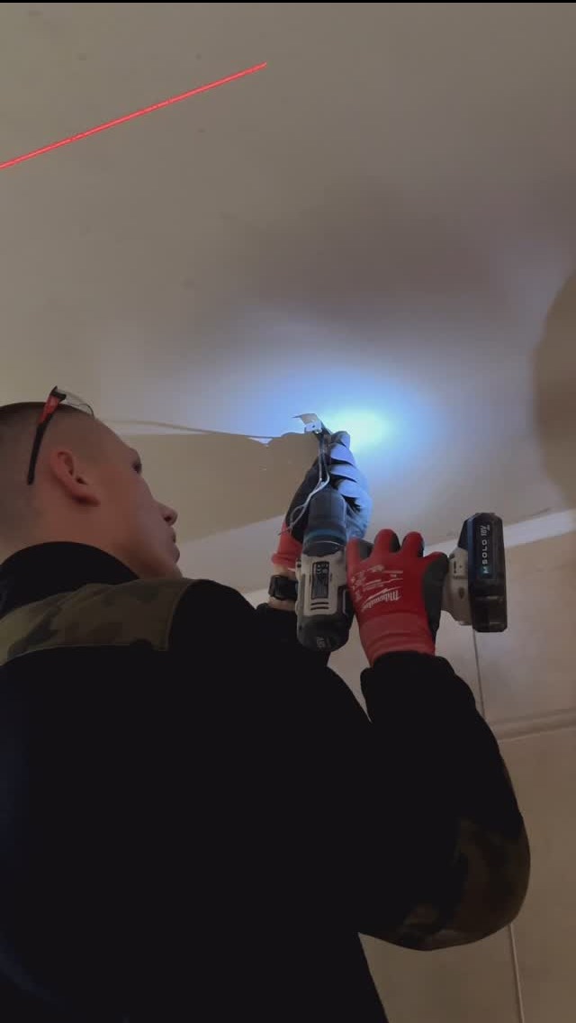 Elektryk w czerwonych rękawicach wierci otwór w suficie, używając wiertarki akumulatorowej Milwaukee, z widoczną wiązką lasera wyznaczającą linię wiercenia.