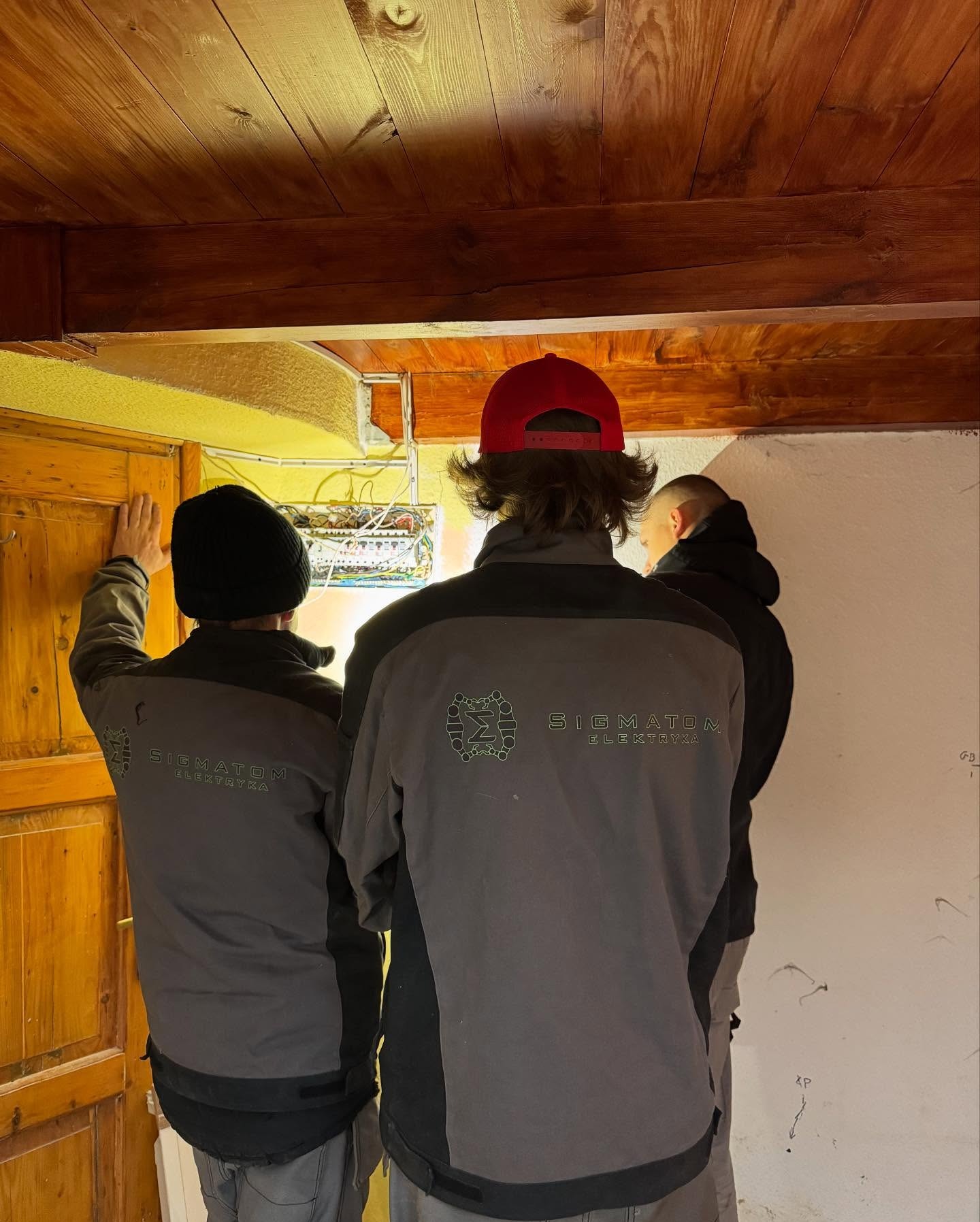 Trzech elektryków w kurtkach firmowych 'SIGMATOM Elektryka' sprawdza skrzynkę elektryczną w budynku z drewnianym sufitem i żółtą ścianą.