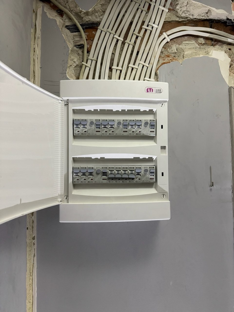 Otwarta skrzynka bezpiecznikowa ETI zamontowana na szarej ścianie, z widocznymi przewodami elektrycznymi w peszlach wchodzącymi w ścianę z ubytkami tynku.