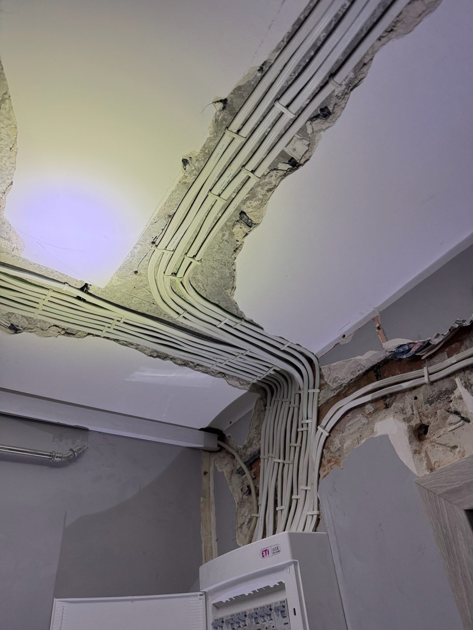 Wiązka białych kabli elektrycznych prowadzonych w bruzdach w suficie i ścianie, z widoczną skrzynką rozdzielczą ETI na dole, w trakcie prac instalacyjnych.