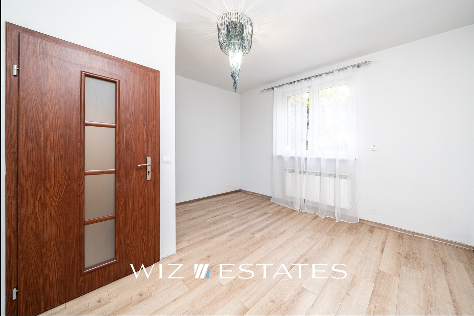 Puste, jasne pomieszczenie z drewnianą podłogą, białymi ścianami, oknem z białymi firankami, nowoczesnym żyrandolem i mahoniowymi drzwiami z przeszkleniami.
