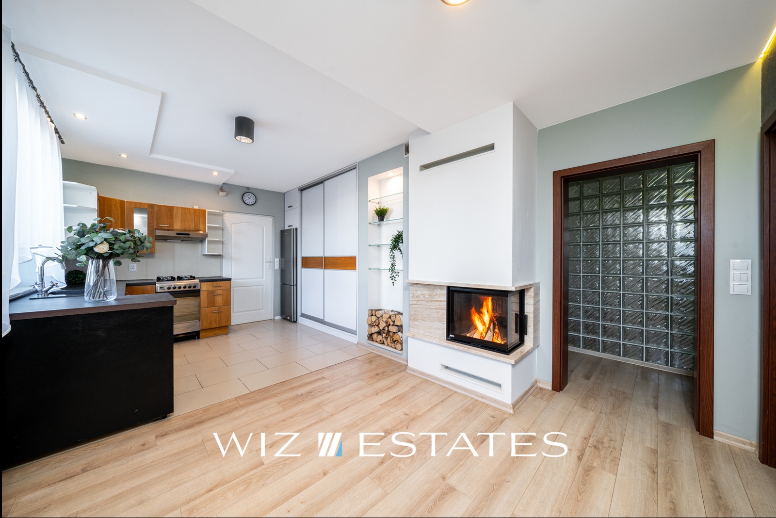 Jasne wnętrze mieszkania z kominkiem, drewnianą podłogą, kuchnią z drewnianymi szafkami i ścianą z luksferów w drzwiach.