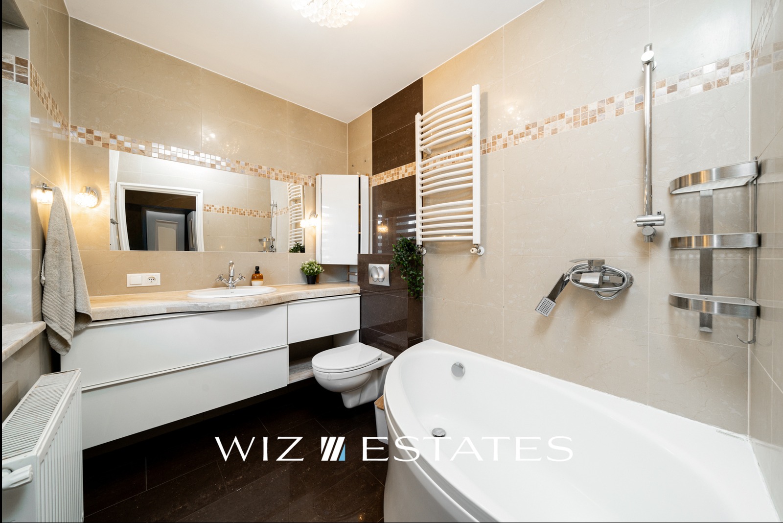 Elegancka łazienka z beżowymi płytkami, białą wanną wolnostojącą, umywalką wpuszczaną w blat i grzejnikiem drabinkowym, oświetlona kryształowym plafonem.