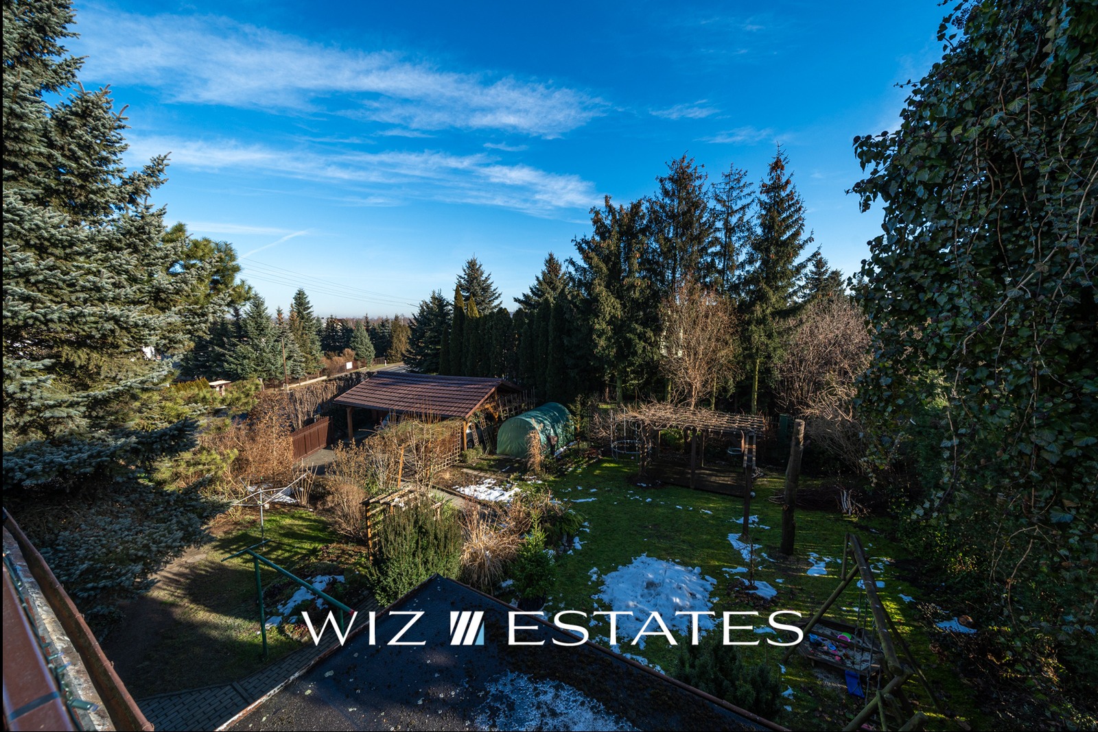 Widok z góry na duży, zadbany ogród w Krakowie z domkiem narzędziowym, szklarnią i pergolą, częściowo pokryty śniegiem, widziany w słoneczny dzień.