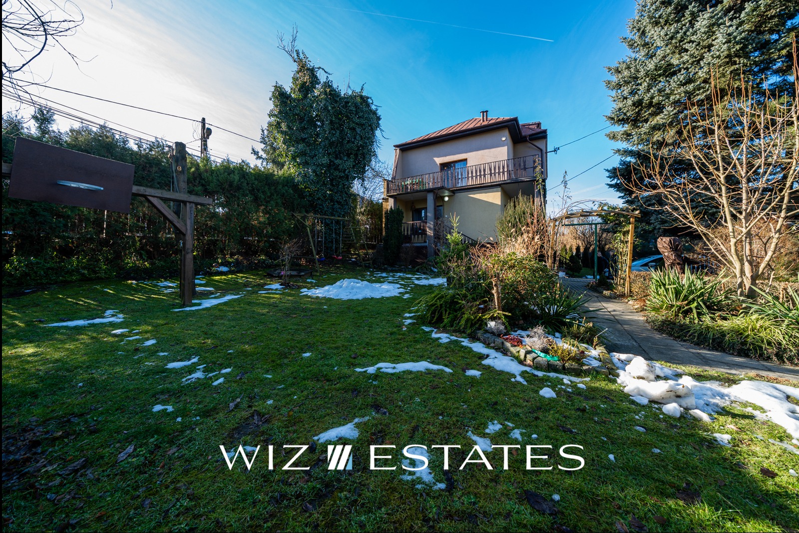 Dwupiętrowy dom z balkonem i zielonym ogrodem pokrytym resztkami śniegu, widoczny kosz do koszykówki i huśtawka.