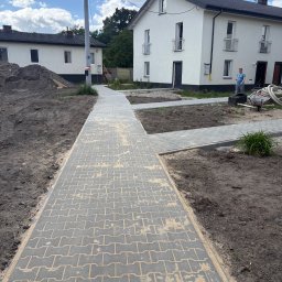 Simply Bruk - Ułożona szara kostka brukowa na posesji przed domem, widok z perspektywy chodnika. W tle budynki mieszkalne i osoba przy pracy.