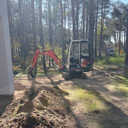 Simply-Bruk - Mała koparka Kubota KX41-3V w lesie, przygotowuje teren pod budowę. Widoczny wykop i sterta ziemi. W tle drzewa i samochód.