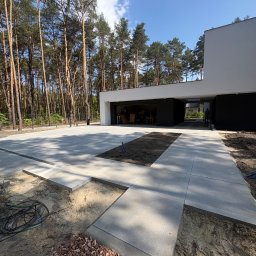 Simply-Bruk - Nowoczesny podjazd z szarych płyt betonowych w trakcie realizacji, widoczne przygotowane podłoże i otoczenie domu z lasem w tle. Częściowo ułożona nawierzchnia.