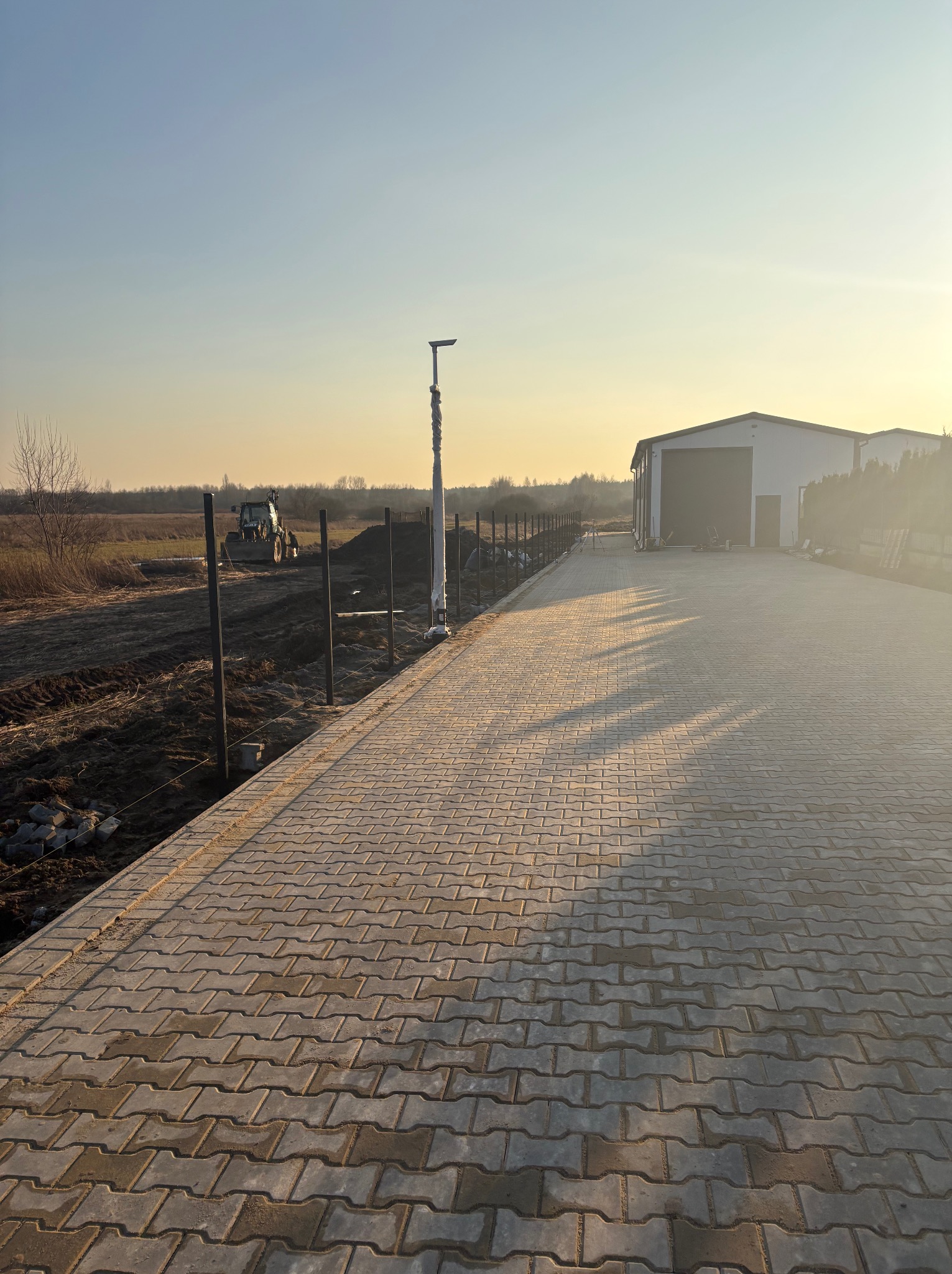 Nowa, szara kostka brukowa ułożona wzdłuż ogrodzenia z metalowych słupków, w tle traktor i budynek magazynowy w porannym słońcu.