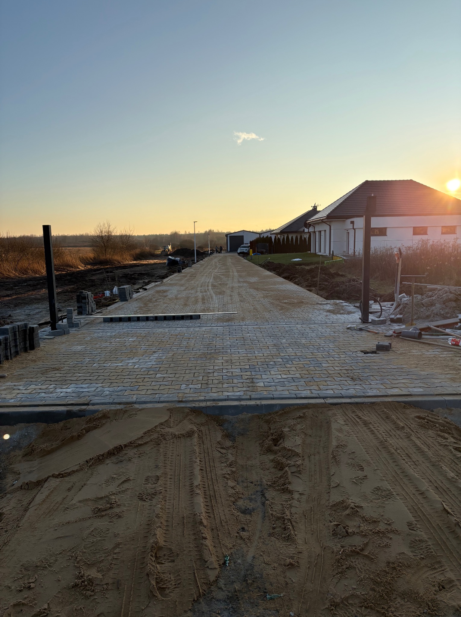 Układanie beżowej kostki brukowej na podjeździe do domu jednorodzinnego, widoczne ślady opon na piasku, w tle zachodzące słońce.