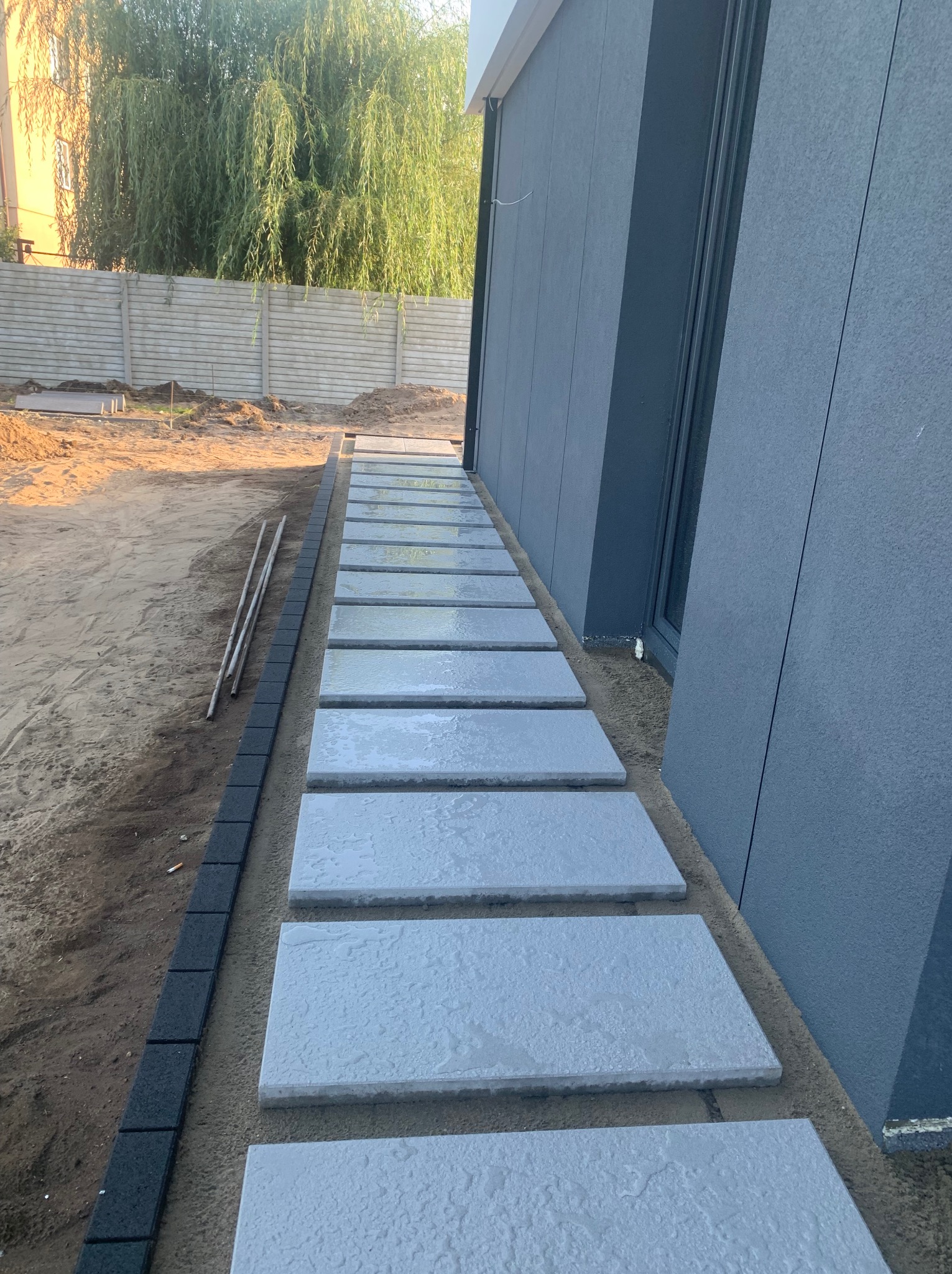 Ścieżka z jasnoszarych płyt betonowych ułożona wzdłuż ściany budynku z szarą elewacją, obrzeże z ciemnych kostek, w tle ziemia, trawa i betonowe ogrodzenie.