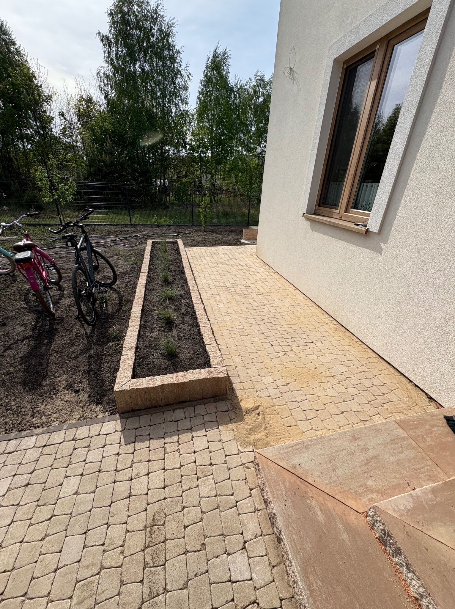 Jasna kostka brukowa ułożona przy domu z oknem, obok rabata z roślinami i dwa rowery. Widok na fragment posesji w słoneczny dzień, z perspektywy ziemi.