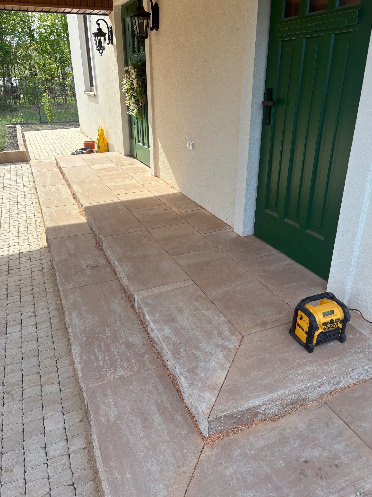 Ułożone schody z kostki brukowej prowadzące do drzwi wejściowych domu. Widoczna kostka brukowa na podjeździe i radio budowlane DeWalt. Detal wykończenia.