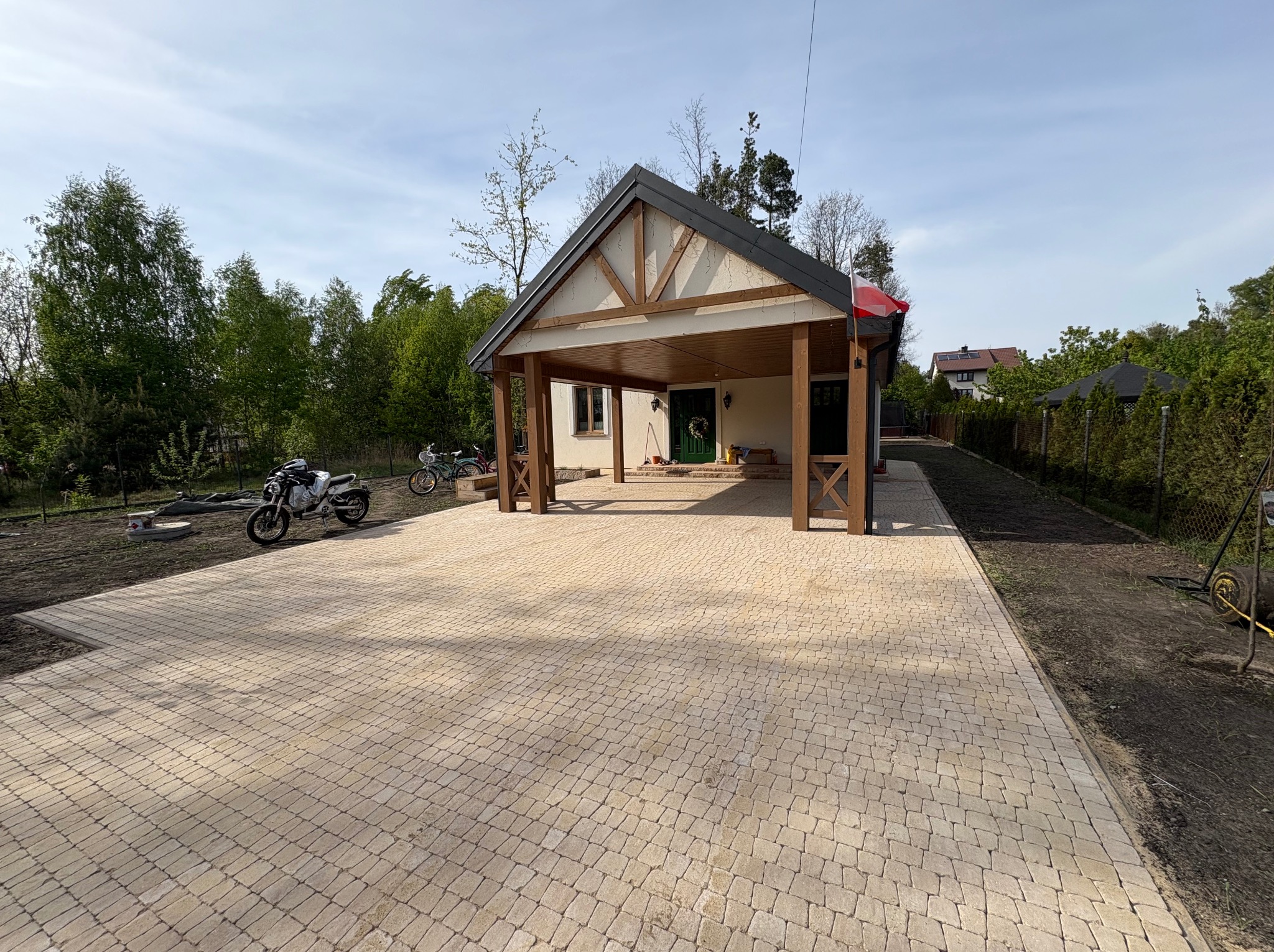 Beżowa kostka brukowa na podjeździe z wiatą, motocykle w tle. Widok na dom z zielonymi drzwiami i wiankiem. Świeżo obsiana ziemia po bokach.