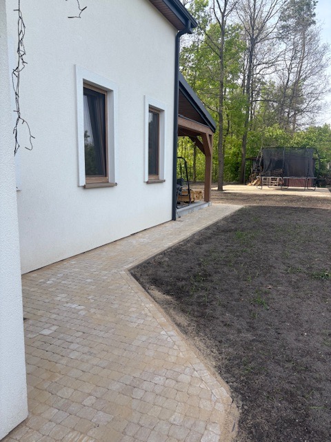 Jasna kostka brukowa tworzy ścieżkę wzdłuż białego domu z drewnianą werandą. W tle widoczne drzewa i trampolina.