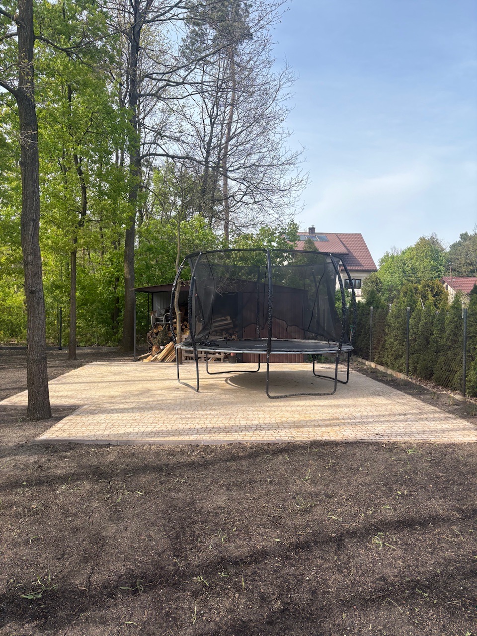 Ułożona kostka brukowa w ogrodzie, tworząca podłoże pod trampolinę. W tle drzewa i budynek z czerwoną dachówką, widoczne w słoneczny dzień.