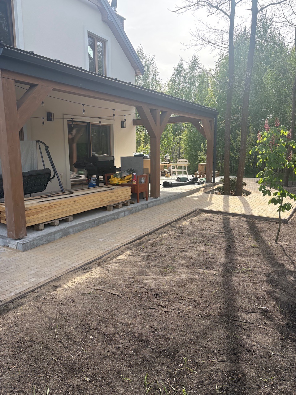 Jasna kostka brukowa tworzy ścieżkę wokół domu z drewnianą pergolą. Widoczny przygotowany teren pod dalsze prace. W tle zielony las.