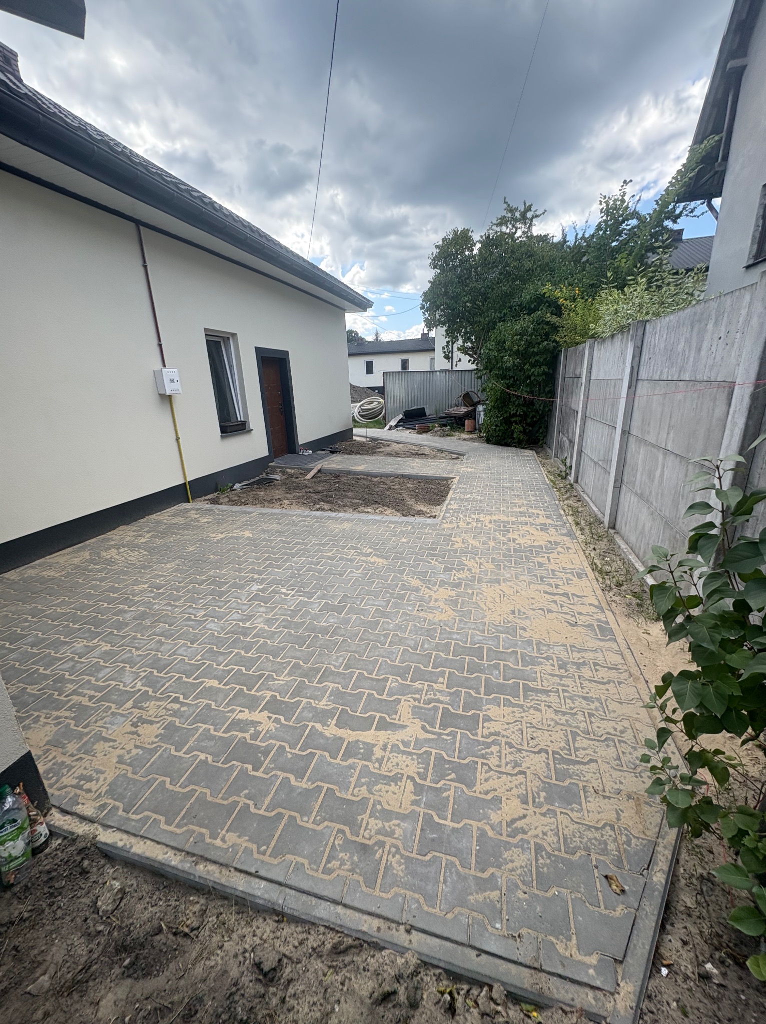 Szara kostka brukowa ułożona na posesji, z fragmentem terenu przygotowanego pod dalsze prace. Betonowy płot w tle, obok budynku z oknem i brązowymi drzwiami.