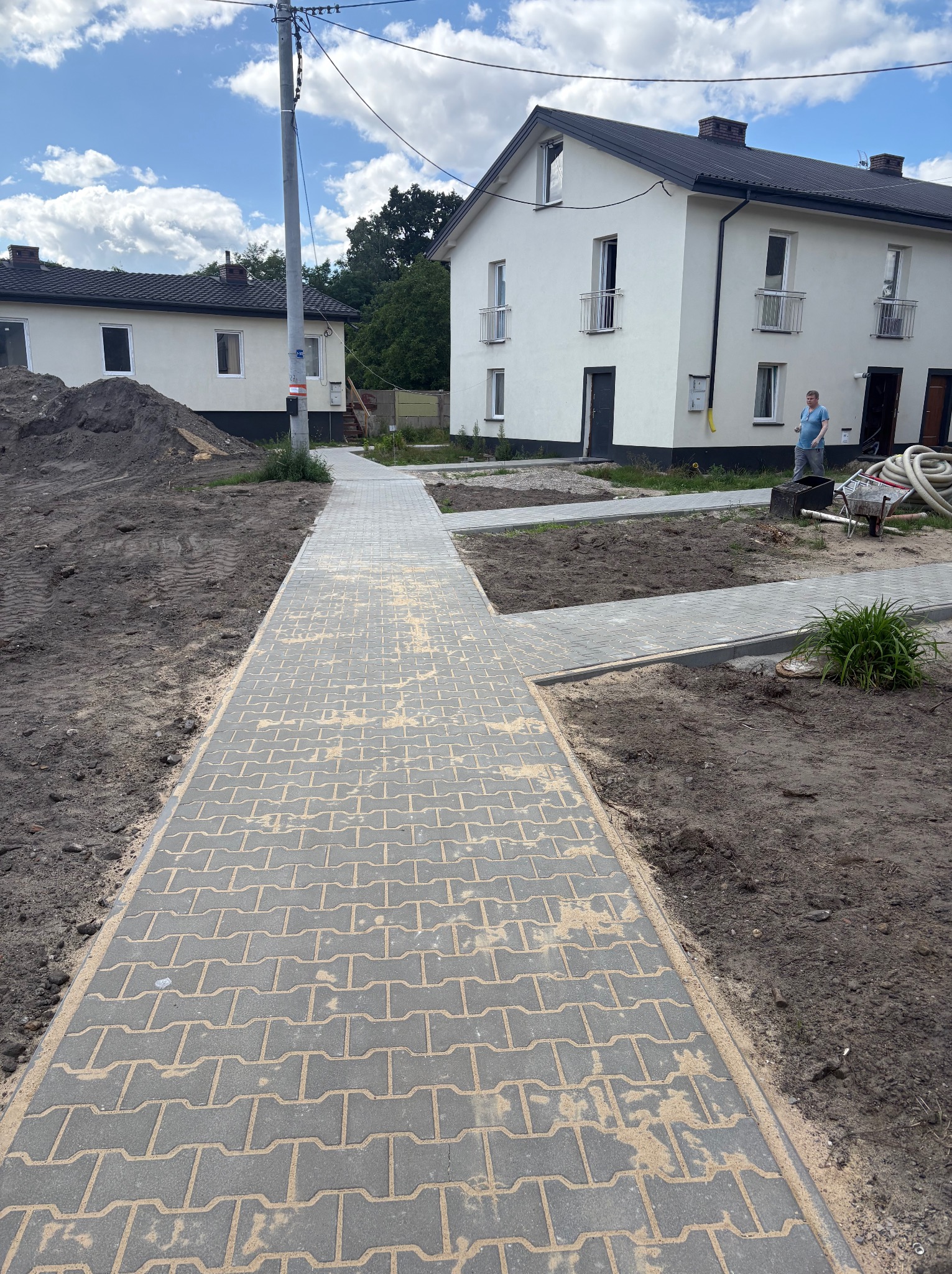 Ułożona szara kostka brukowa na posesji przed domem, widok z perspektywy chodnika. W tle budynki mieszkalne i osoba przy pracy.