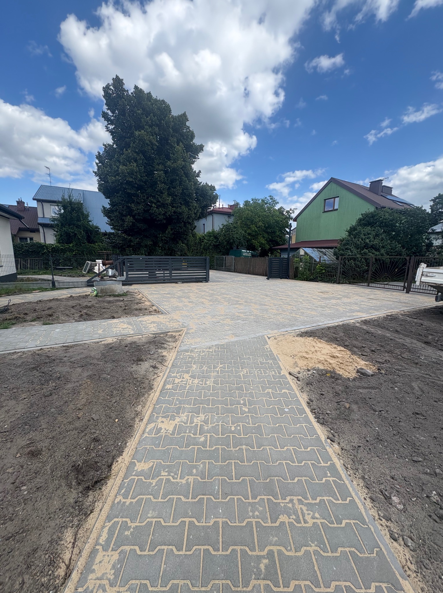 Szara kostka brukowa ułożona na podjeździe i chodniku, z widocznym fragmentem metalowego ogrodzenia i furtki. W tle zieleń drzew i zabudowa jednorodzinna w słoneczny dzień.