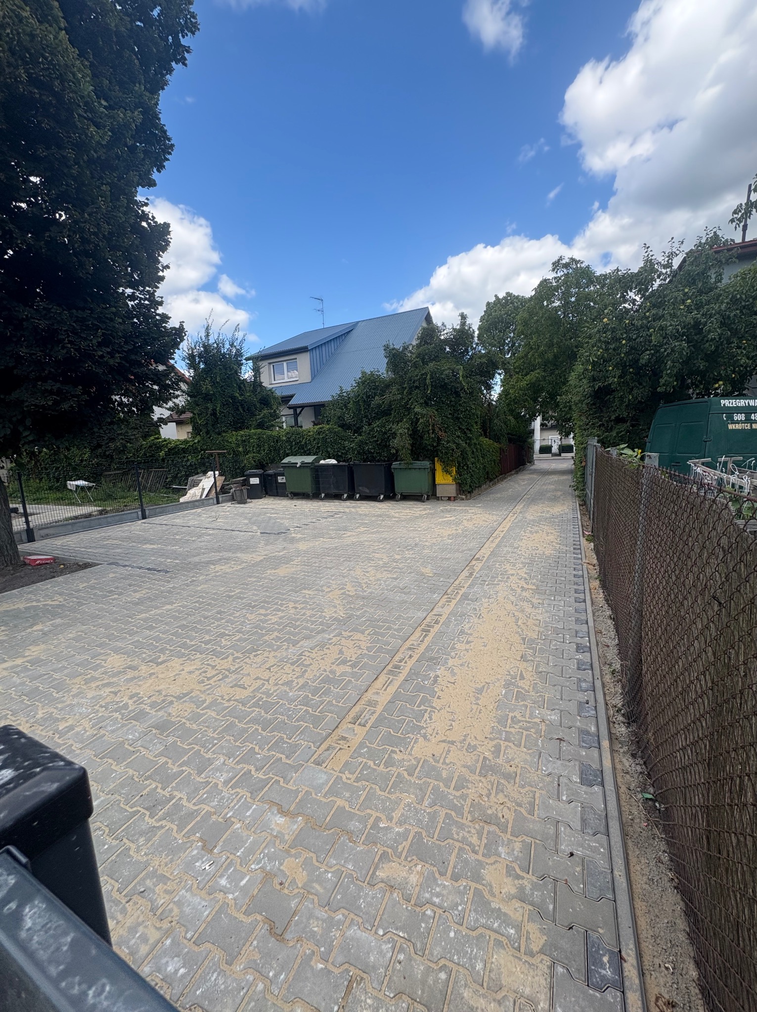 Świeżo ułożona, szara kostka brukowa na podjeździe, z piaskiem w spoinach. Widoczne ogrodzenie z siatki i zieleń drzew. Błękitne niebo z chmurami w tle.