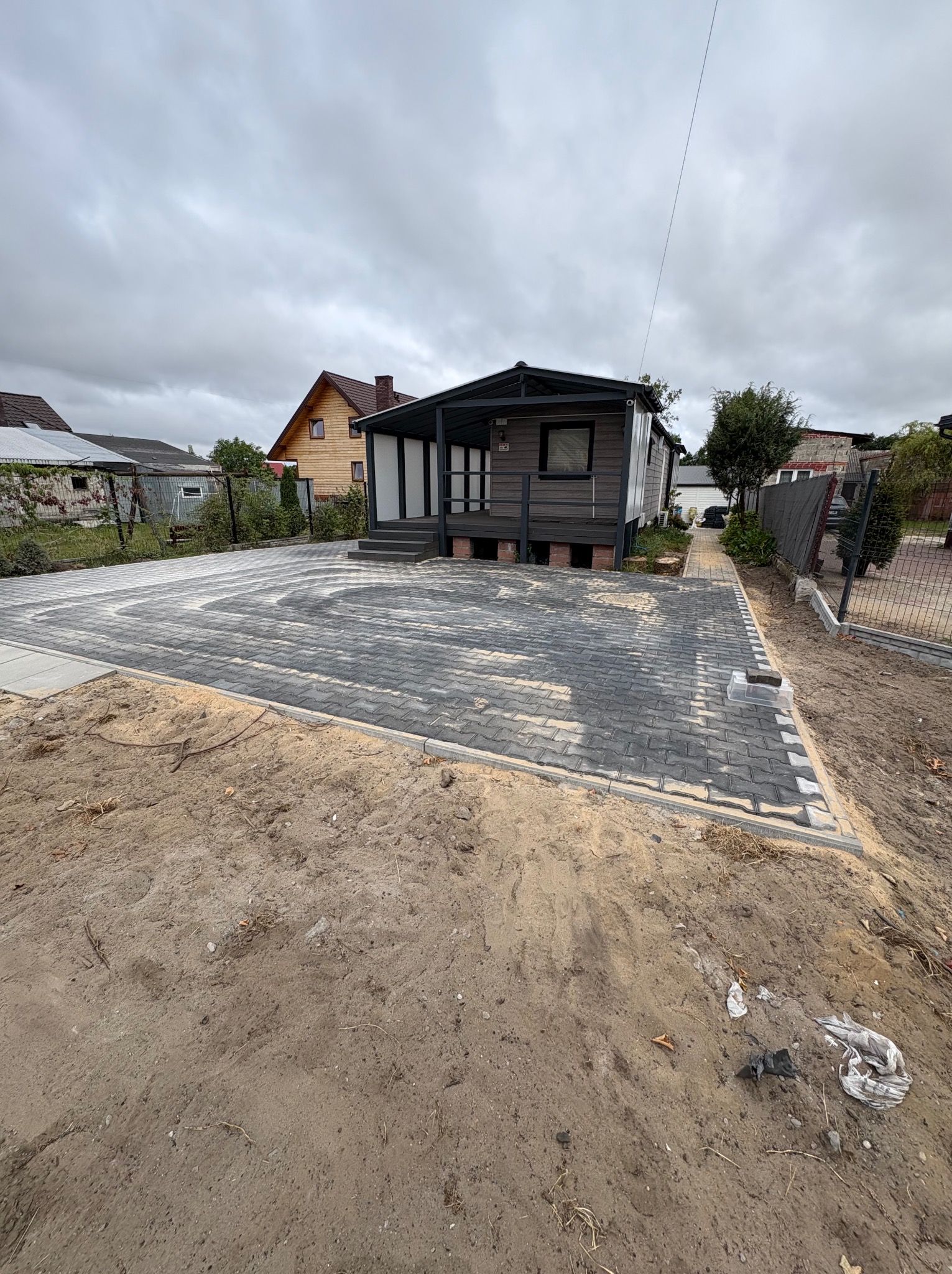 Nowa, szara kostka brukowa ułożona na podjeździe przed domem z szarymi ścianami i czarnym dachem. Widoczne obrzeża i piasek. Pochmurne niebo w tle.