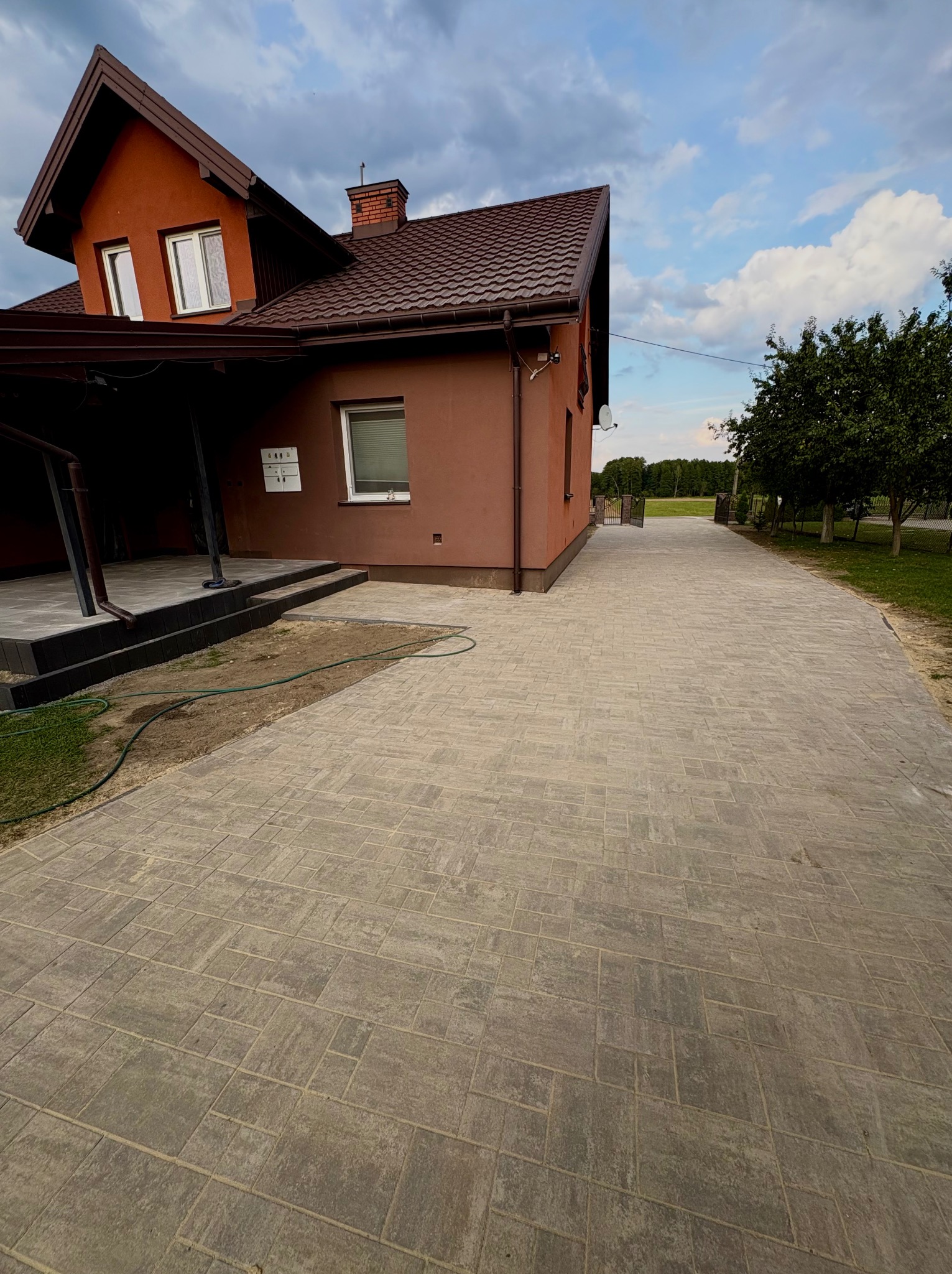 Ułożona kostka brukowa prowadząca do domu z brązowym dachem i elewacją. Widoczny fragment ogrodu i niebo z chmurami.