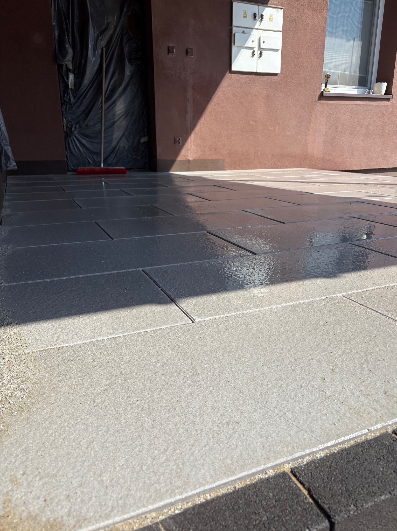 Świeżo ułożona, dwukolorowa kostka brukowa przed budynkiem. Część kostki jest jeszcze mokra, co sugeruje niedawne czyszczenie lub deszcz. Widoczny fragment elewacji.