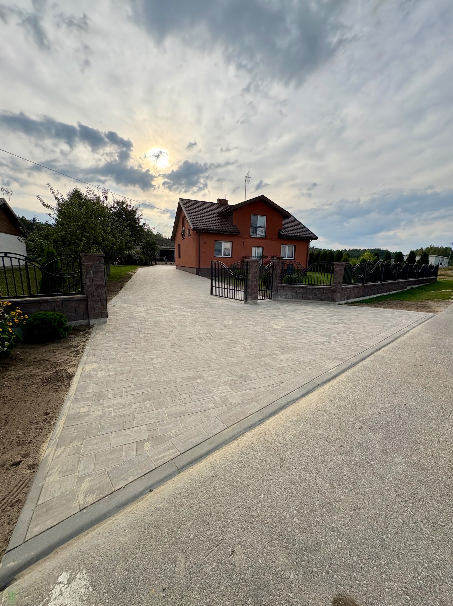 Jasna kostka brukowa prowadzi do domu z czerwonej cegły, ogrodzenie z metalowymi przęsłami i ceglanymi słupkami po bokach. Niebo pochmurne.