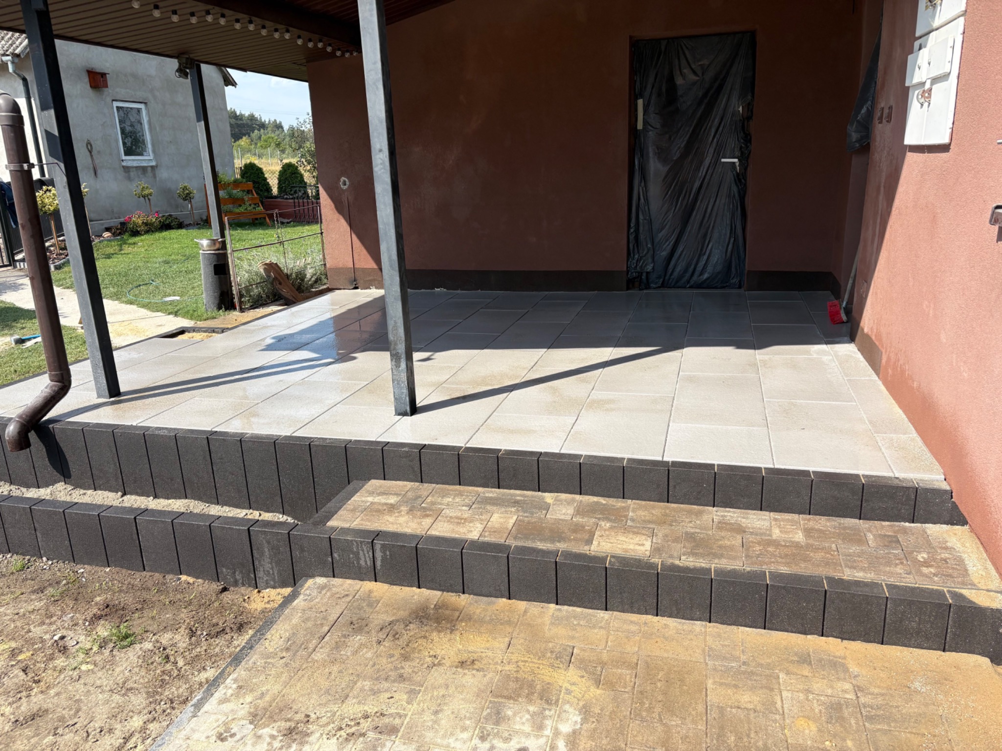 Wykończony taras z jasnych płyt na podwyższeniu, obrzeża z ciemnej kostki. Widoczny fragment domu z zadaszeniem i ogrodem w tle.