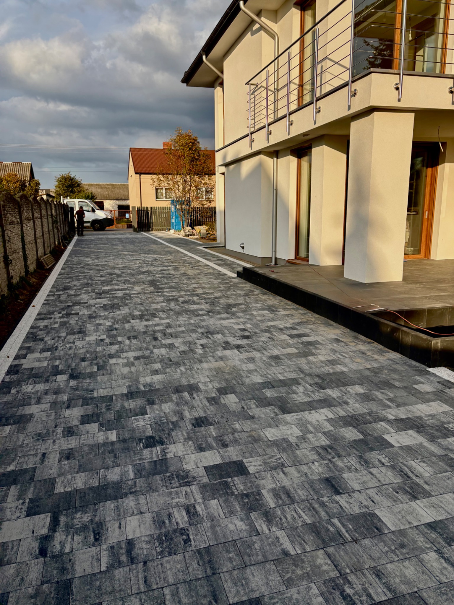 Ułożona, szara kostka brukowa prowadząca do domu z balkonem, widoczny fragment ogrodzenia i osoba w oddali. Nowoczesny design, długa, równa powierzchnia.