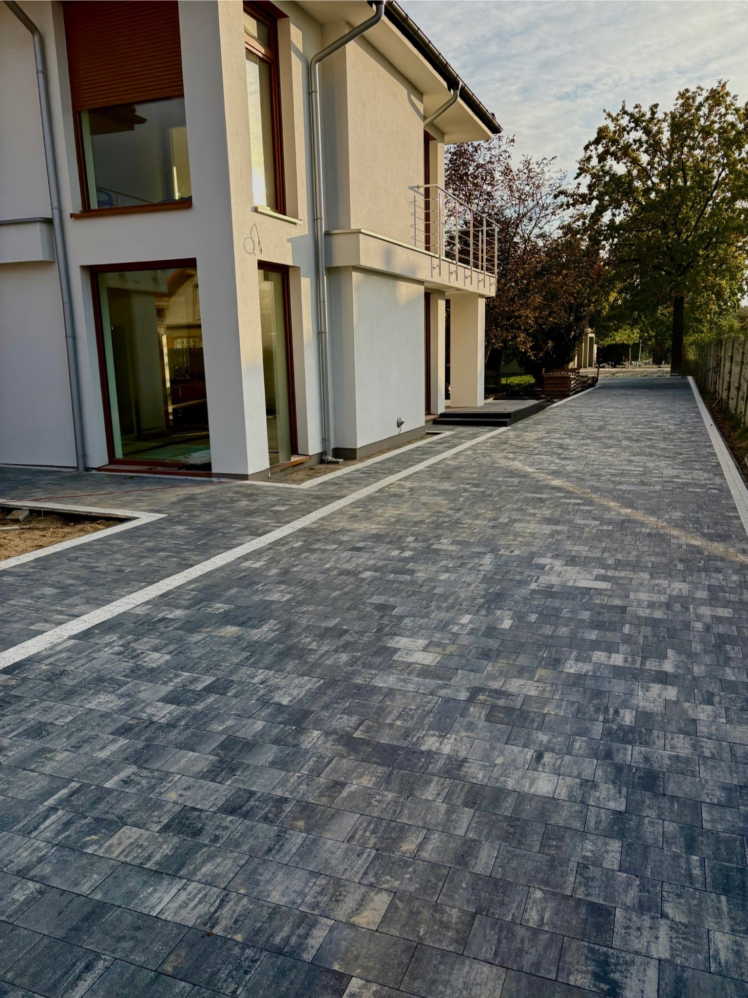 Nowoczesna posesja z szarą kostką brukową ułożoną w geometryczny wzór, obrzeża z jasnego kamienia. Widok na biały dom z balkonem i duże okna.