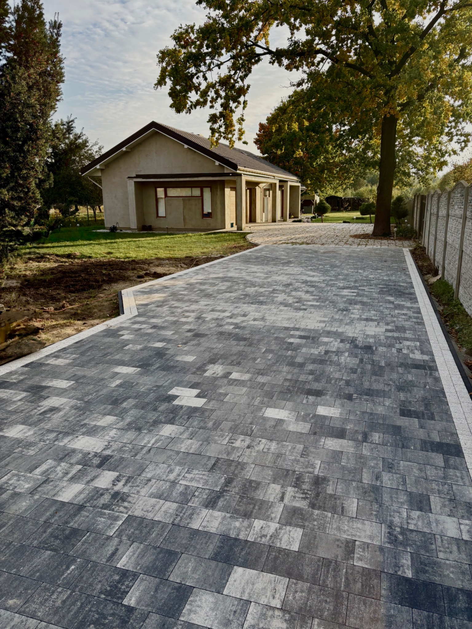 Szeroki podjazd z szarej kostki brukowej prowadzący do domu, obramowany białym obrzeżem, z betonowym ogrodzeniem w tle i zielenią. Perspektywa z dołu.