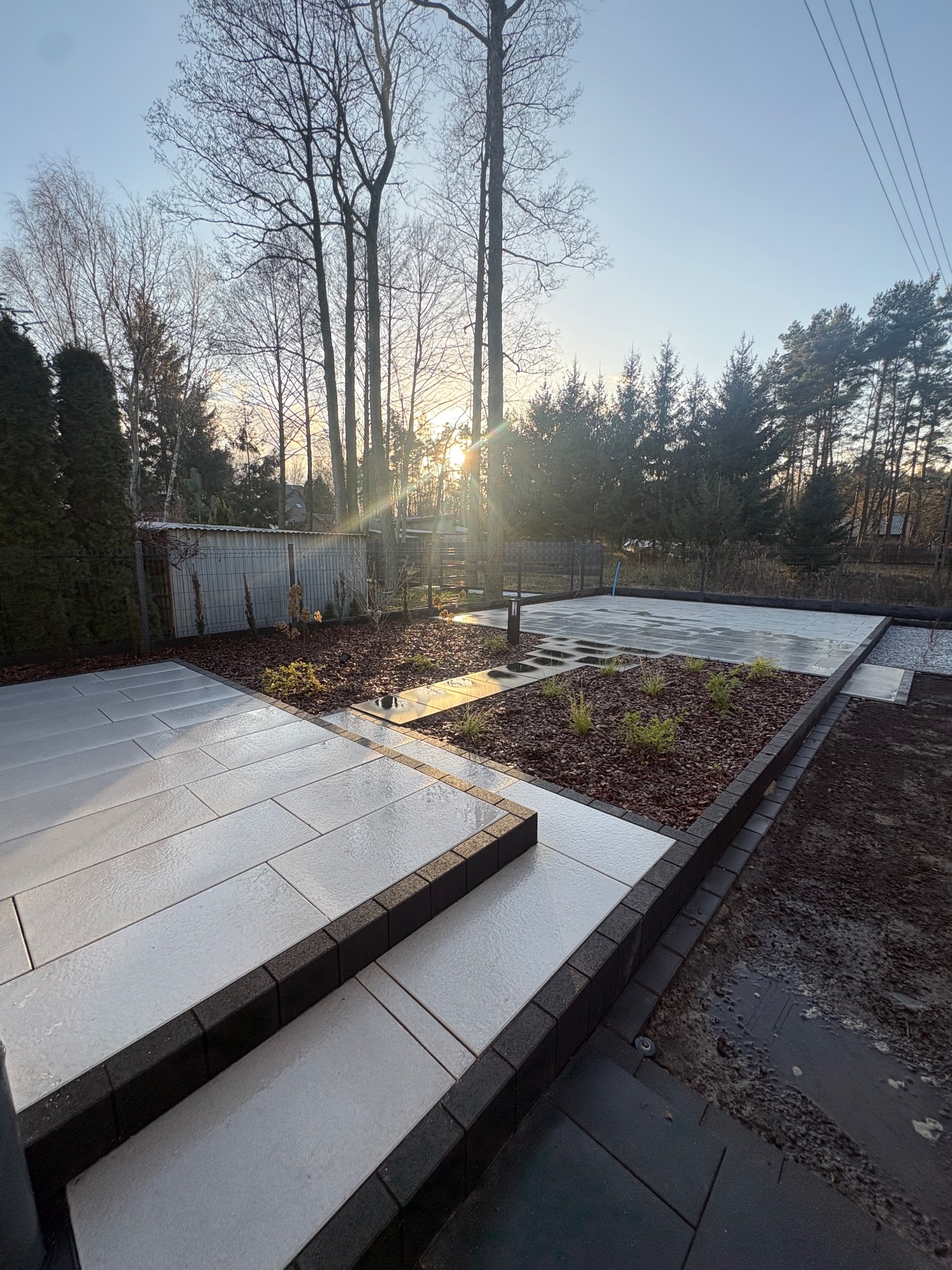 Nowoczesny taras z jasnoszarej kostki brukowej, obrzeża z ciemnej kostki, rabaty z korą i roślinami. W tle drzewa i zachodzące słońce.
