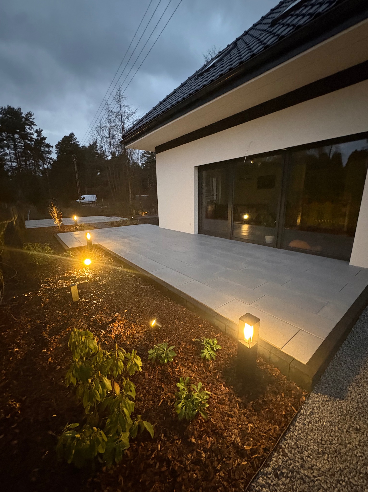 Oświetlony taras z szarych płyt, obrzeża z kostki, rabata z korą i lampami ogrodowymi. Nowoczesny dom z dużymi oknami przesuwnymi w tle, wieczorny krajobraz.