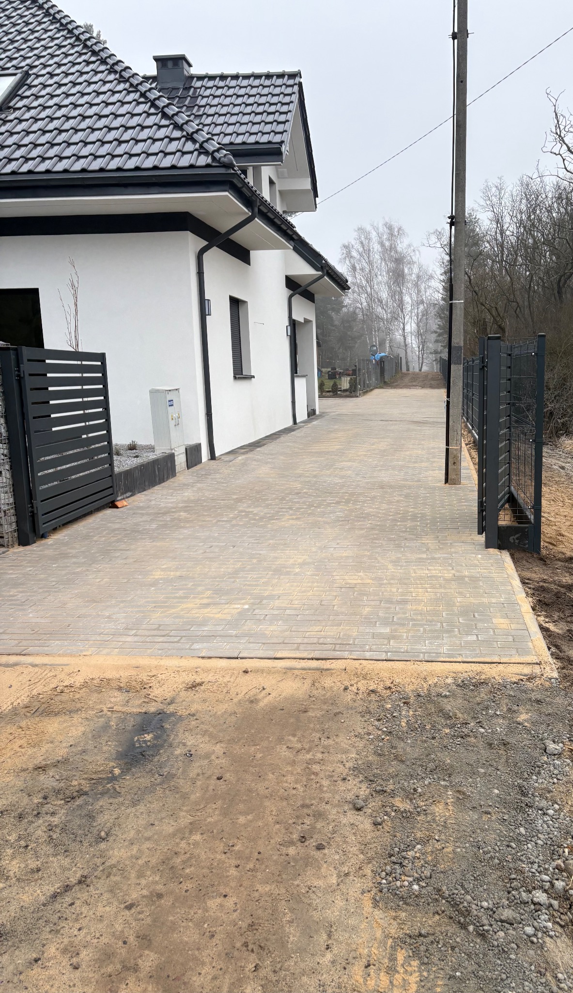 Nowoczesny podjazd z kostki brukowej w odcieniach szarości, prowadzący do białego domu z czarnym dachem. Widoczne metalowe ogrodzenie i przygotowany teren pod dalsze prace.