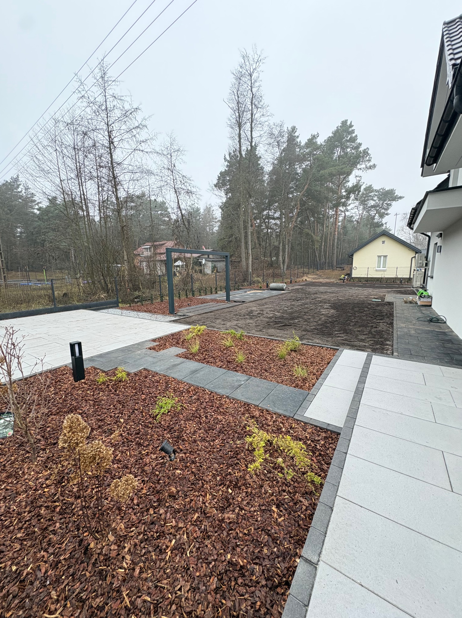 Nowoczesny ogród z jasną kostką brukową, rabatami z korą i minimalistyczną pergolą w tle. Widoczny fragment domu i drzewa. Pochmurna pogoda.