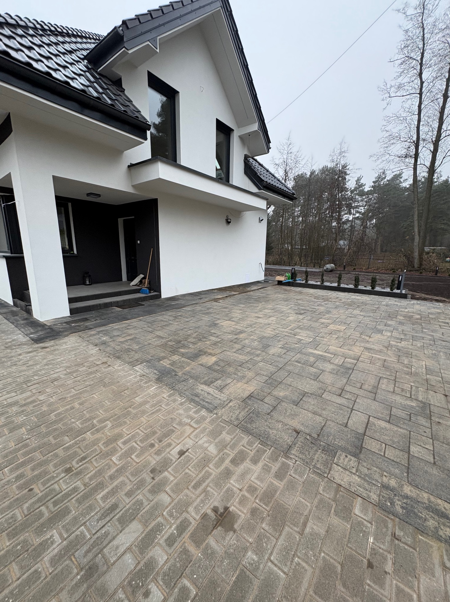 Nowoczesny dom z szarą kostką brukową na podjeździe. Widoczne obrzeża i świeżo posadzone tuje. Minimalistyczny styl budynku i otoczenia.