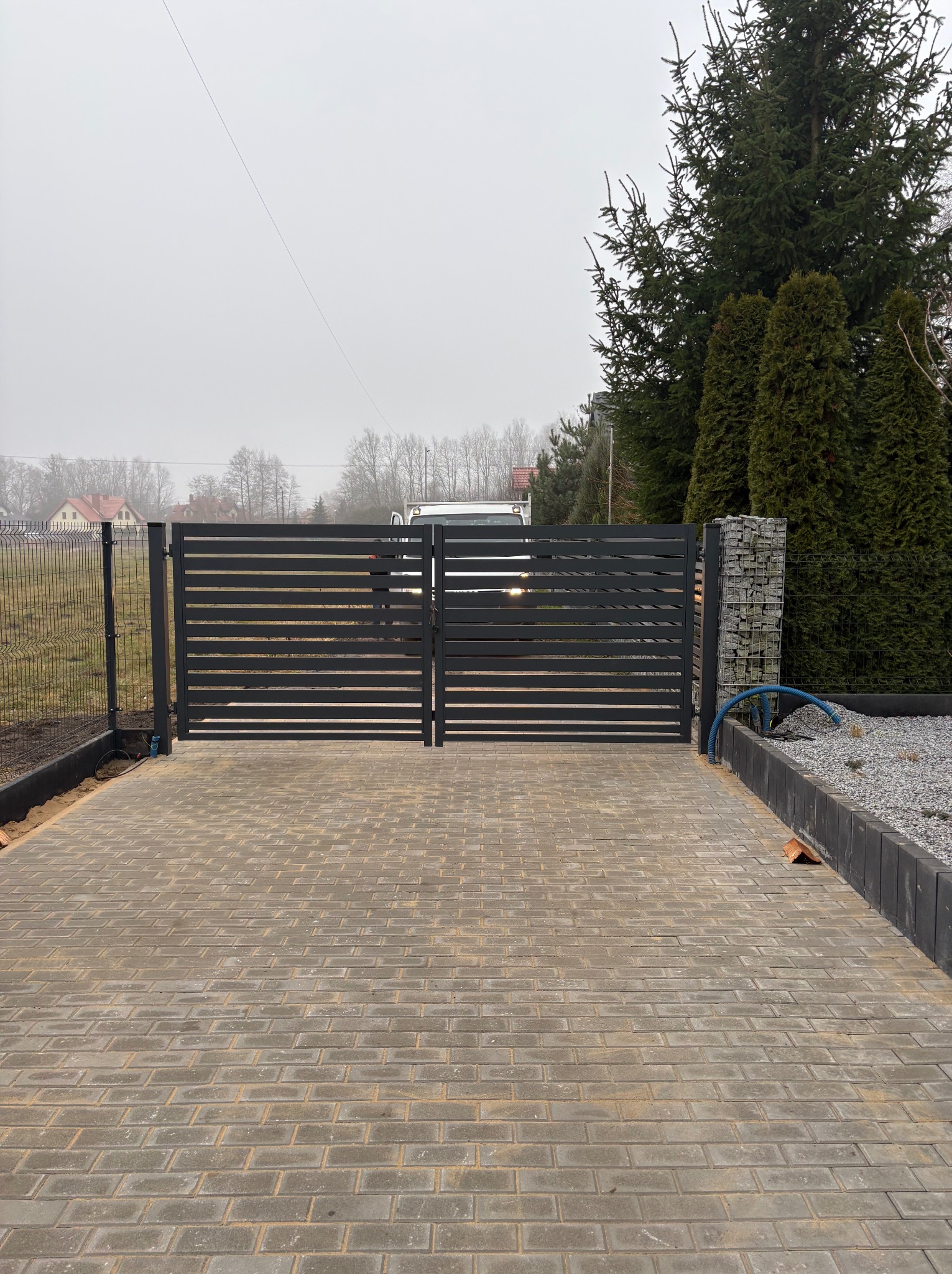 Nowoczesna, pozioma brama wjazdowa z szarej kostki brukowej i gabionowym słupkiem. Widoczne ogrodzenie panelowe i zieleń w tle. Pochmurny dzień.