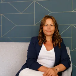 Portret kobiety w granatowej marynarce siedzącej na szarym fotelu na tle ściany z geometrycznym wzorem.