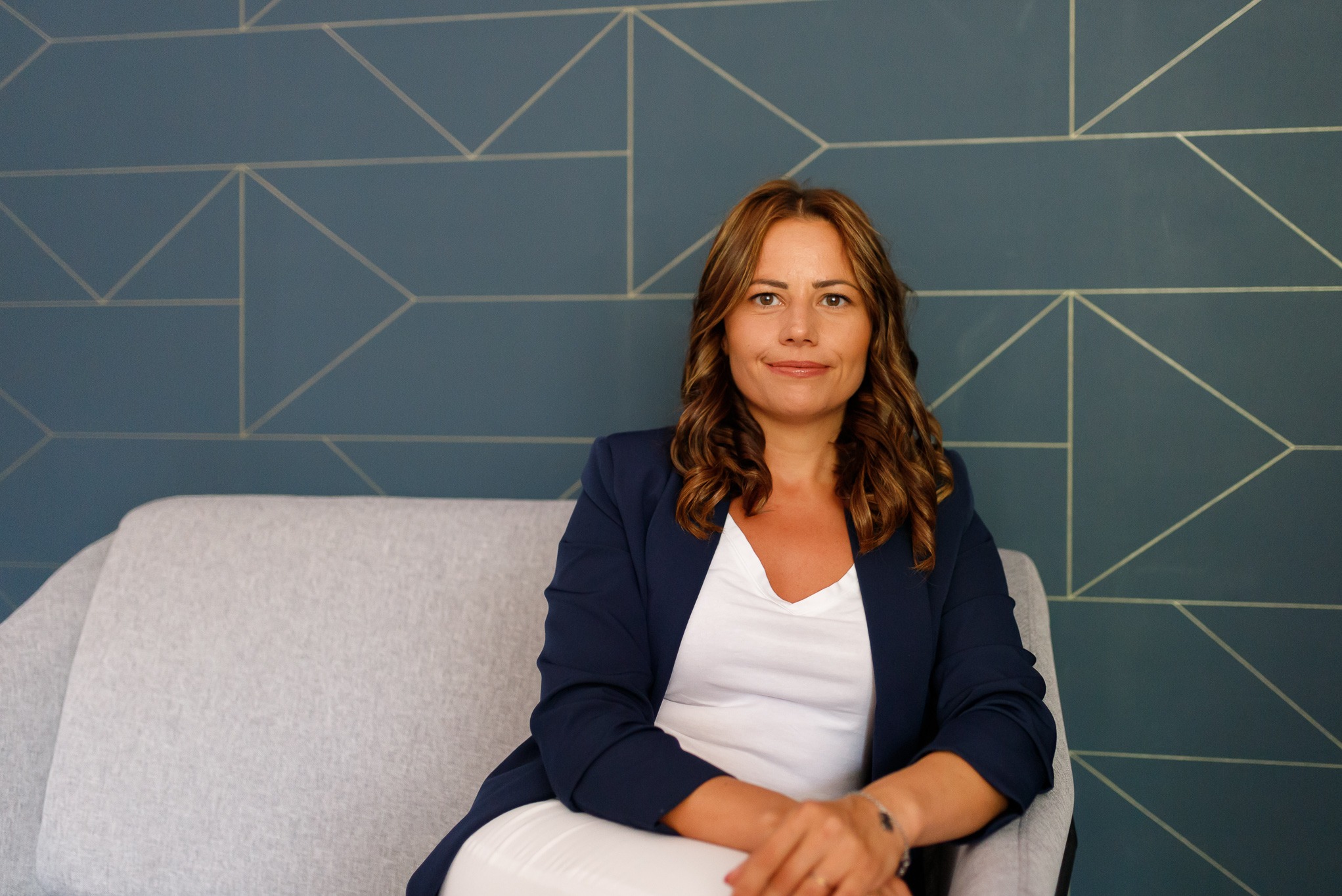 Portret kobiety w granatowej marynarce siedzącej na szarym fotelu na tle ściany z geometrycznym wzorem.